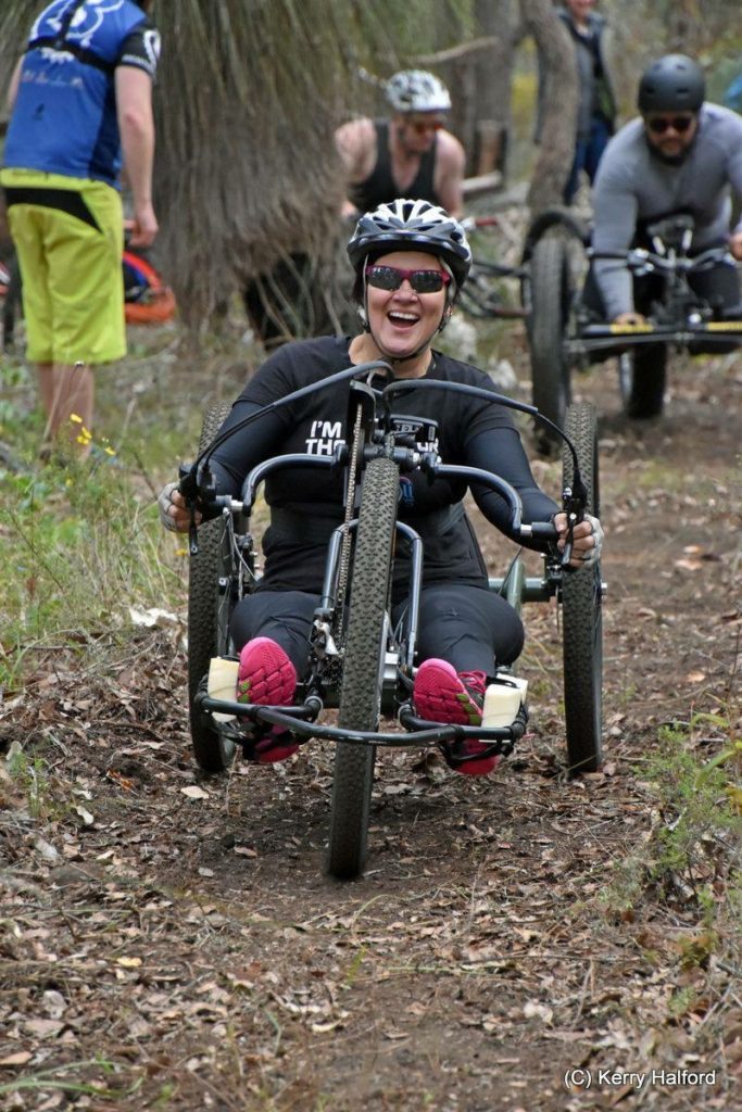 a woman is in a handcycle riding in the forest. she is smiling broadly.