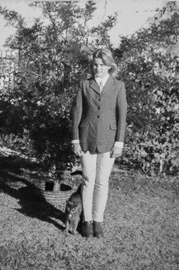 a young woman is standing up straight, she is wearing a horseriding kit of jodphurs, a riding jacket and shirt 