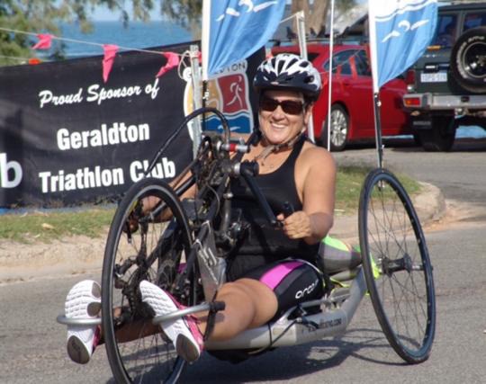 a woman is on a adapted handcycle. She is in front of a sign that says geraldton triathlon club.