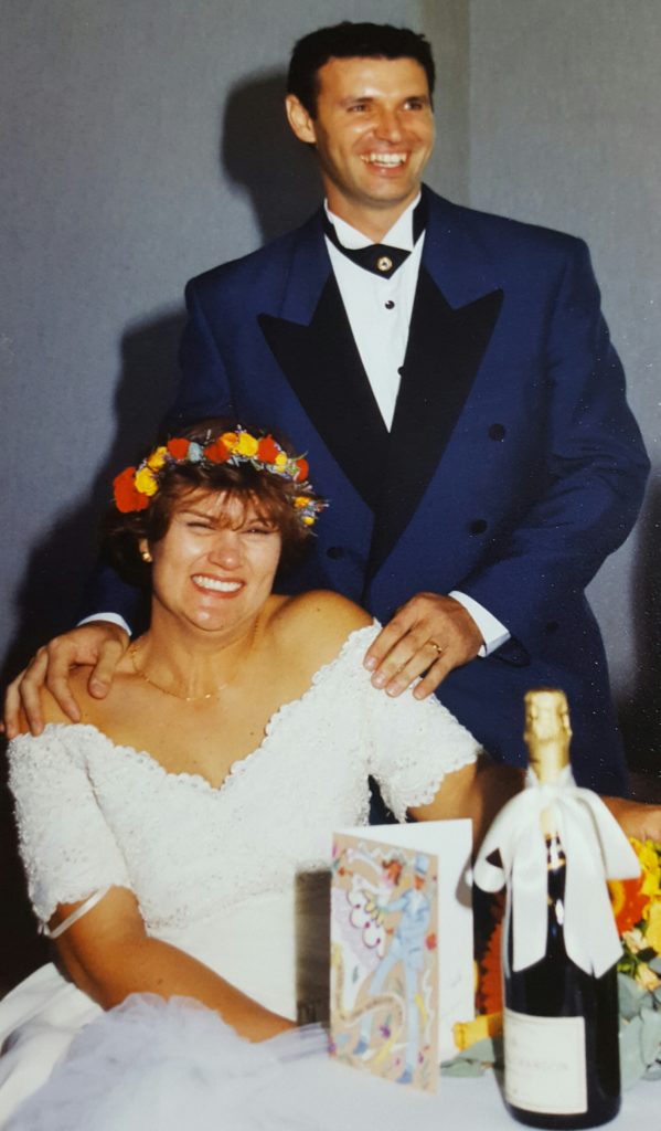 a wedding photo, the groom is standing behind his wife, he wears a blue suit and is smiling. The bride is sitting and smiling. She wears a crown of bright flowers and an off the shoulder wedding dress.