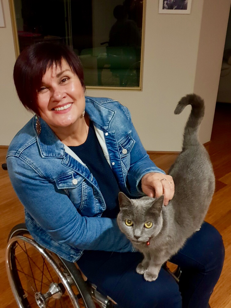 a woman with brown hair is sitting in a wheelchair. She is wearing a denim jacket, is smiling, and has a grey cat on her lap.