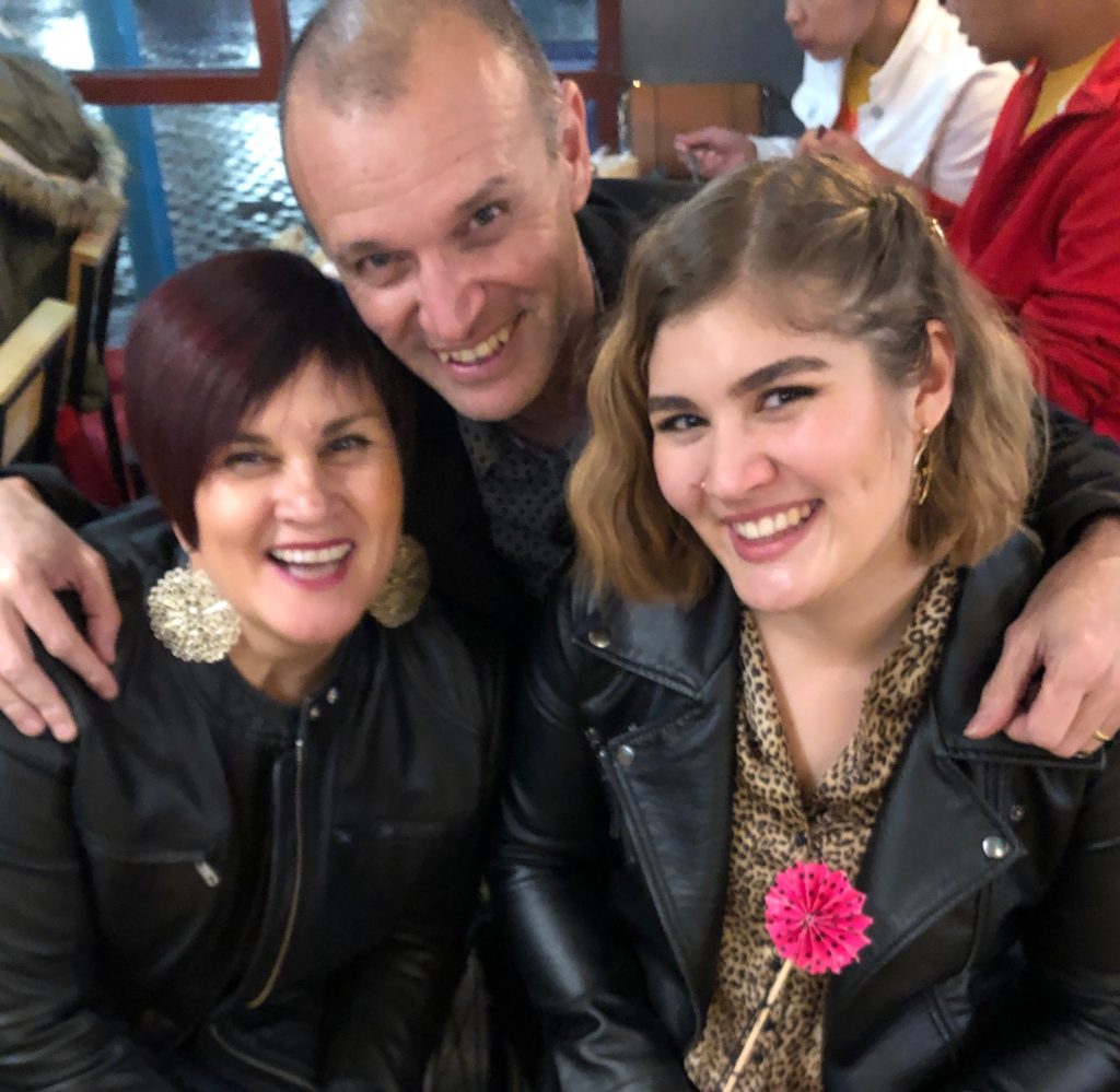 a man stands between two women, his arms are around them both. They are all smiling.