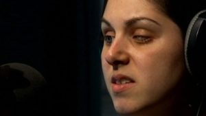 a headshot of woman wearing headphones speaking into a microphone. The woman has one eye open and one eye partially closed.