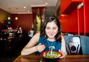 a woman with black hair is about to eat a mouthful of food, she is in a restaurant and has a smile on her face. She is wearing a blue sleeveless dress