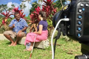 a man in a floral shirt and a woman in a pink skirt and blue t shirt sit next to each other on a grass area in front of some tropical plants with bright red flowers. A camera lense is visible in the foreground.