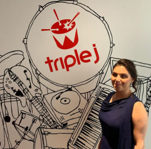 a woman is standing in front of a sign that says Triple J. She is wearing a blue sleeveless dress and is smiling at the the camera