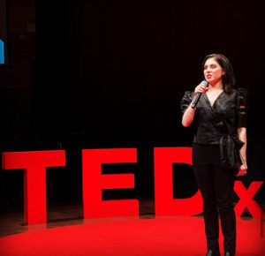 a woman in standing on a stage in front of a red sign that says TED x . She is wearing a black shirt and pants and is speaking into a microphone