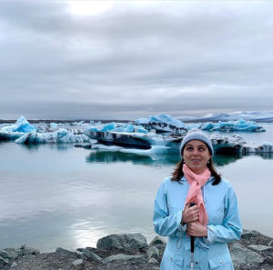 an image of a woman standing in front of an ice pack. she is wearing light blue coat and beanie and a pink scarf. she is holding a cane in front of her and smiling at the camera