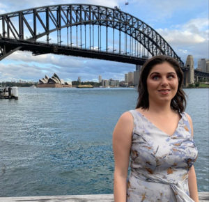 A woman is wearing a light blue dress, she has black hair, she is smiling at the camera and the sydney harbour bridge is in the background