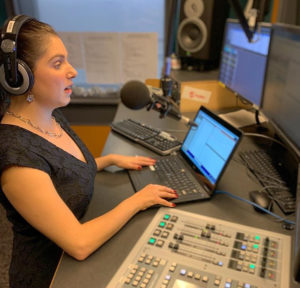 a woman wearing headphones is sitting in front of a desk with a keyboard and a microphone. It is in a recording studio.