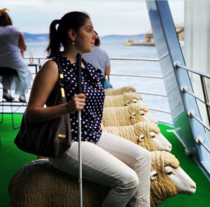 a woman wearing a polka dot shirt and white pants sits on a deck of a ship on a seat shaped like a sheep. it is one of a row of sheep statues lined up on the deck looking out to sea. She is holding a white cane by her side.